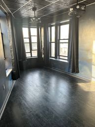 Empty urban loft-style room with black painted walls and dark hardwood floor, three large windows with black curtains letting in sunlight and a city street view, plus industrial ceiling fans.