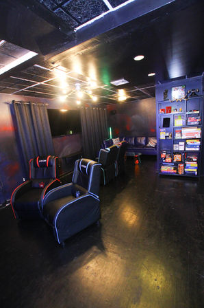 Modern home game room/basement media room with black ceiling, row of black gaming recliners facing a wall-mounted TV, leather sofa and a tall shelf stacked with board games under warm ambient lighting.
