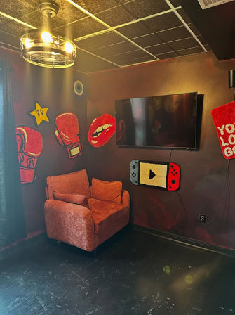 Cozy gaming/media nook with rust-colored armchair, wall-mounted TV, dark accent wall decorated with playful pop-art rugs (boxing gloves, star, lips, handheld game controller) and an industrial ceiling light.