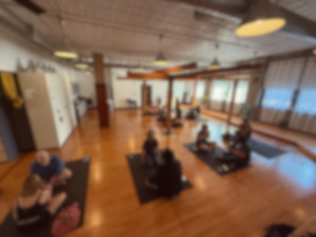 Loft-style group yoga class on black mats in a bright studio with hardwood floors, wall mirrors and hanging pendant lights.
