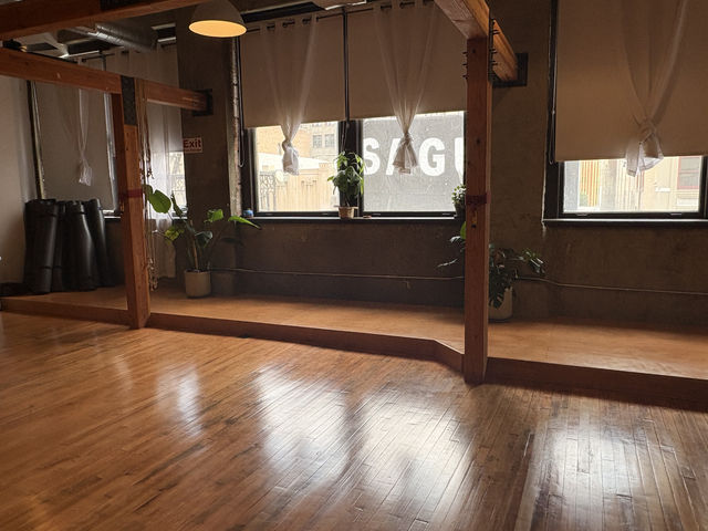 Sunlit urban loft studio with polished hardwood floors, wooden beams, large windows with tied shades, potted plants and stacked yoga mats.