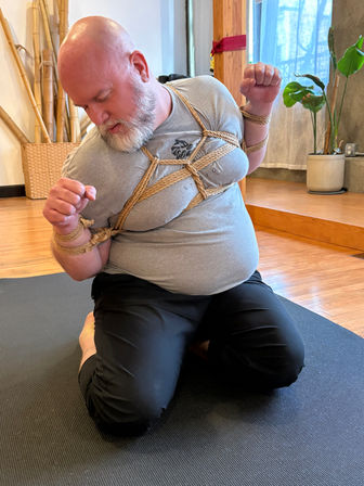 Bearded bald man kneeling on a black yoga mat in a bright studio, wearing a gray T‑shirt and black pants, bound in decorative natural‑fiber rope chest harness and wrist ties, wooden floor, bamboo decor and potted plants in the background