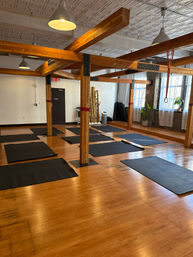 Sunlit loft-style yoga studio with exposed wooden beams and hardwood floors, black exercise mats spaced on the floor, hanging pendant lights, large windows, plants and red suspension straps.