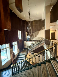 Historic theater lobby stairwell with carpeted steps, ornate iron-and-brass railing, dramatic hanging stage lights on ropes, tall windows and vertical wooden acoustic panels bathed in warm light.