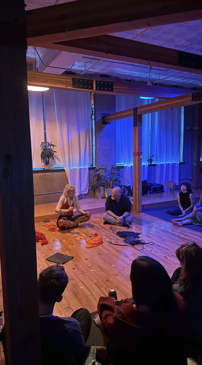 Loft-style wellness studio with exposed wooden beams and blue-purple mood lighting, people seated in a circle on a hardwood floor listening to a woman leading a seated yoga/meditation workshop with plants and sheer curtains in the background.