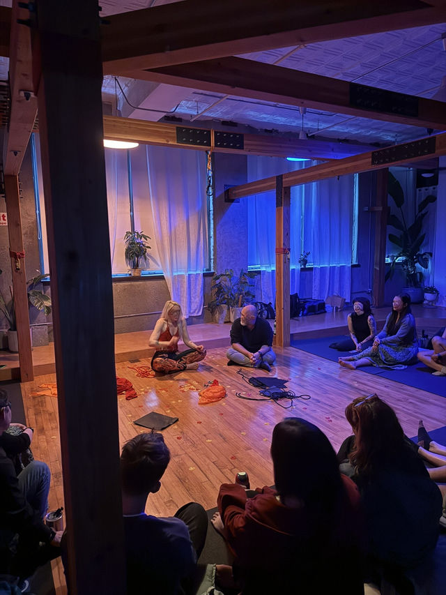 Loft-style wellness studio with exposed wooden beams and blue-purple mood lighting, people seated in a circle on a hardwood floor listening to a woman leading a seated yoga/meditation workshop with plants and sheer curtains in the background.