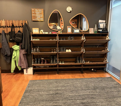 Modern office entryway with wooden coat hooks and hanging jackets, multiple open shoe racks holding boots and sneakers, two teardrop mirrors above a wooden shelf, hardwood floor and gray area rug