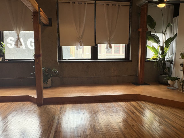 Sunlit urban loft studio interior with polished hardwood floors, raised wooden platform, exposed timber beams, large windows with tied roller shades, and potted plants