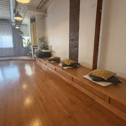 Cozy sunlit loft-style studio with polished hardwood floor, raised wooden platform topped with stacked cushions, exposed brick column, industrial pendant lights and potted plants by tall windows — calm yoga and meditation space