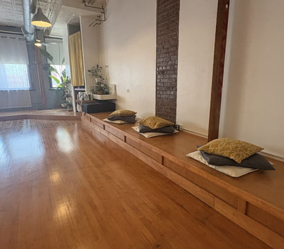 Cozy sunlit loft-style studio with polished hardwood floor, raised wooden platform topped with stacked cushions, exposed brick column, industrial pendant lights and potted plants by tall windows — calm yoga and meditation space