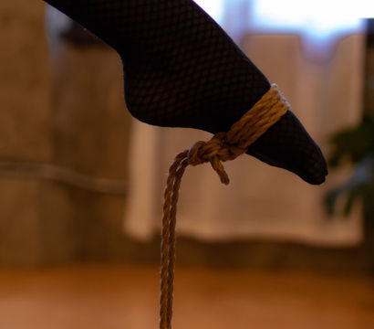 Close-up of an ankle wearing black fishnet stocking with a knotted natural-fiber rope tied around it, warm indoor bokeh background