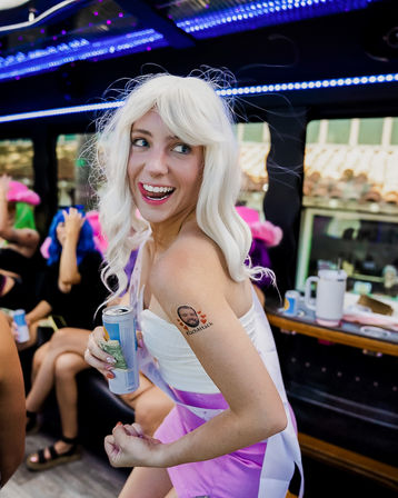 Smiling woman in a white wig and pink outfit inside a LED-lit party bus, holding a canned drink and showing a small temporary portrait tattoo on her upper arm as friends celebrate in the background.