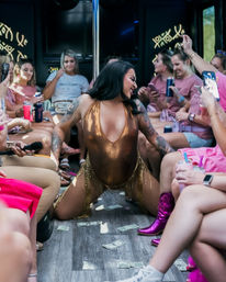 Performer in a gold bodysuit dancing around a pole on a party bus as women in pink cheer, film on phones and toss dollar bills during a bachelorette-style celebration.