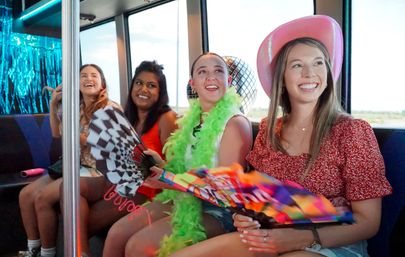 Four smiling women seated in a decorated party bus holding colorful hand fans, a green feather boa, disco ball, and a pink cowboy hat.