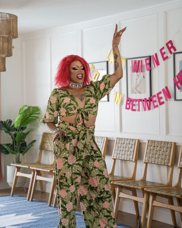 Drag performer with bright red wig and green floral tie-front top and matching wide-leg pants striking a lively pose in a modern living room decorated with a pink party banner and woven wooden chairs