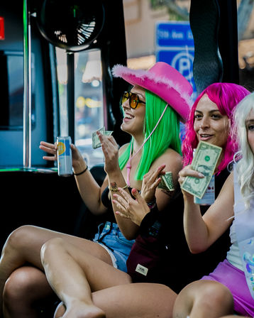 Three people in colorful wigs and summer outfits laughing inside a city party bus, holding dollar bills and drinks — pink cowboy hat, neon green and magenta hair, upbeat urban nightlife vibe.