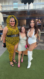 Outdoor bar patio group photo of three smiling people on artificial turf: one in a yellow leopard-print hooded bodysuit with black belt, one in a sparkly mini dress and pink heels, and one in a plunging white bodysuit with fishnets, tattoos and white thigh-high boots.