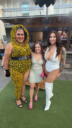 Outdoor bar patio group photo of three smiling people on artificial turf: one in a yellow leopard-print hooded bodysuit with black belt, one in a sparkly mini dress and pink heels, and one in a plunging white bodysuit with fishnets, tattoos and white thigh-high boots.