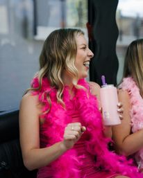 Laughing woman in a bright pink feather boa and dress holding a pink insulated tumbler at a girls' night celebration.