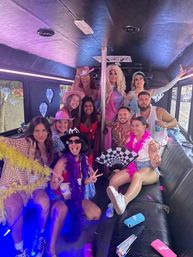 Group of friends and drag performers posing inside a neon-lit party bus around a dance pole, wearing colorful outfits, cowboy hats, feather boas, sunglasses and holding fans and drinks — lively bachelorette-style celebration.