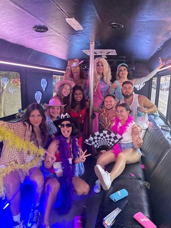 Group of friends and drag performers posing inside a neon-lit party bus around a dance pole, wearing colorful outfits, cowboy hats, feather boas, sunglasses and holding fans and drinks — lively bachelorette-style celebration.
