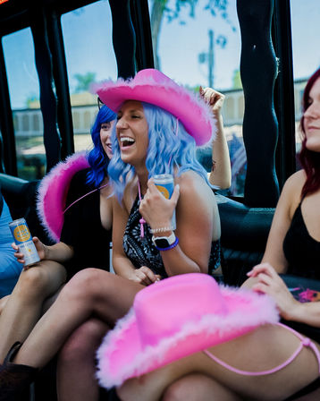 Group of friends laughing inside a party vehicle, woman in a blue wig and fuzzy pink cowboy hat holding a canned drink, playful bachelorette/girls‑night vibe.