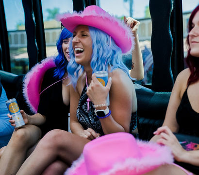 Group of friends laughing inside a party vehicle, woman in a blue wig and fuzzy pink cowboy hat holding a canned drink, playful bachelorette/girls‑night vibe.