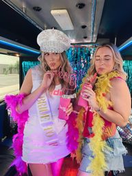 Bachelorette party on a party bus: bride-to-be in a jeweled captain hat and 'Bride to Be' sash sipping from a curly straw, friend in a denim skirt and yellow feather boa holding pink drinks in front of a teal fringe backdrop and neon lights.