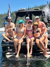 Six friends in swimsuits and sun hats relaxing on a motorboat swim platform, holding drinks and smiling on a sunny mountain lake during a summer boat party