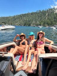 Three friends in bikinis and cowboy hats relaxing on a boat bow during a sunny summer day on a mountain lake, pine-covered shoreline and other boats in the background.