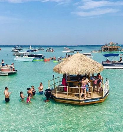 Floating tiki bar in shallow turquoise water with people swimming and socializing, anchored boats nearby under a sunny blue sky.