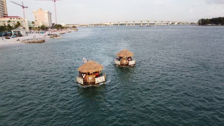 Aerial shot of two floating tiki-hut boats with passengers on turquoise coastal water near a sandy beachfront resort and a long concrete bridge