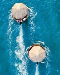 Aerial view of two octagonal thatched-roof tiki huts floating on turquoise ocean, each with an outboard motor leaving white wake trails across the water.