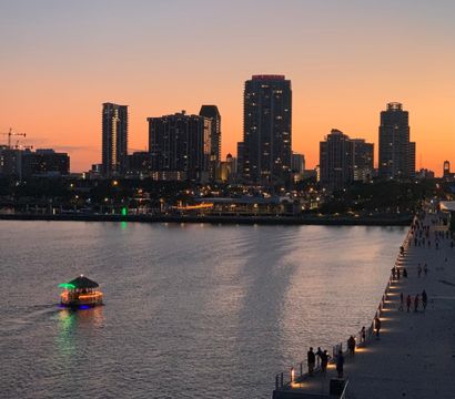 Sunset waterfront city skyline with high-rise silhouettes, a colorful lit boat on calm water, and people strolling along a lit pier promenade.