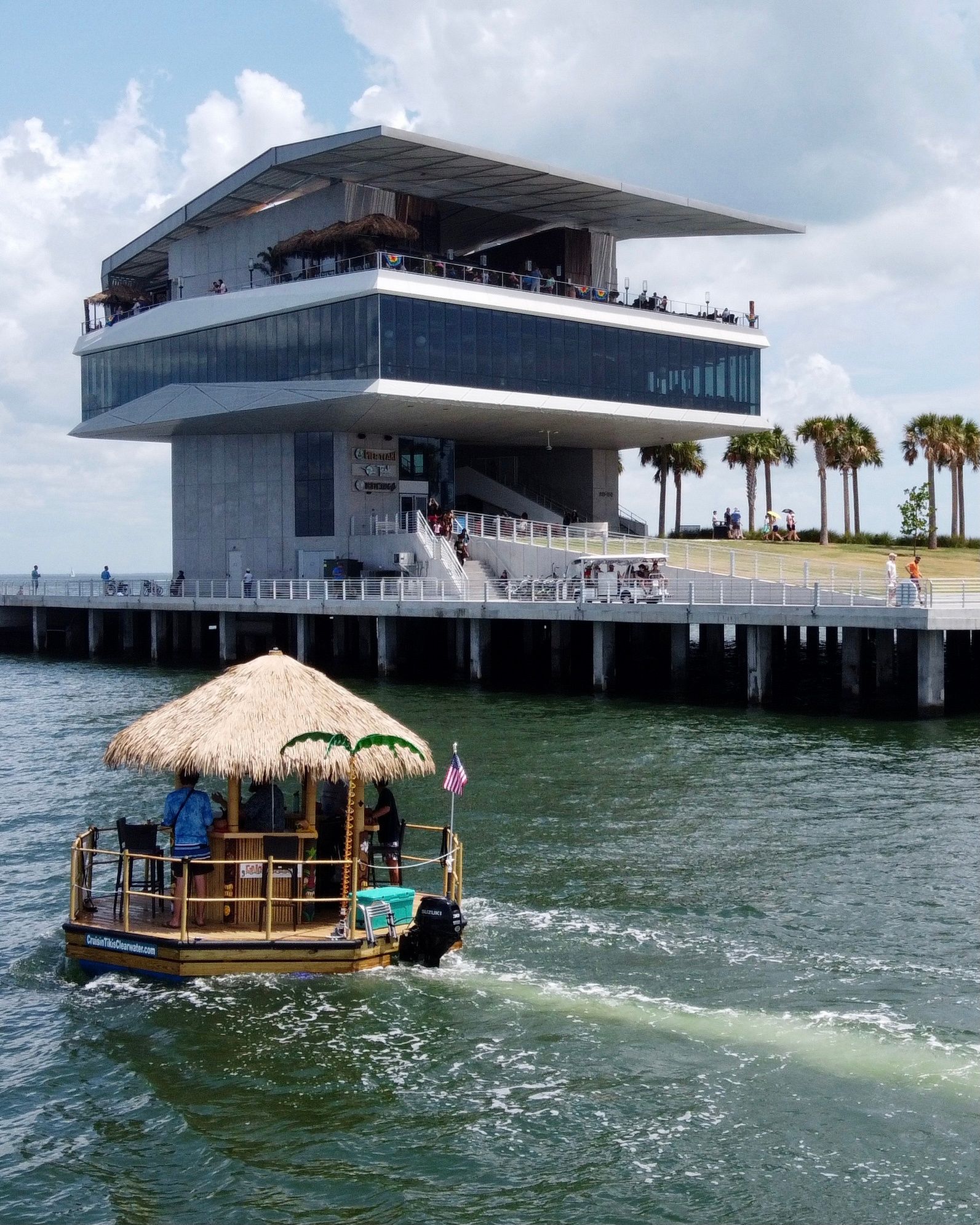 Thatched-roof tiki pontoon boat cruising on a bay past a modern multi-level waterfront observation building and palm-lined pier on a sunny day.