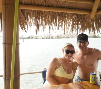 Couple in swimwear smiling under a thatched tiki hut at a waterfront bar, wooden counter with canned drinks and coastal homes and a bridge across the bay visible in the background.
