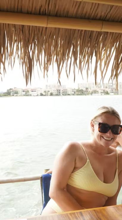 Couple in swimwear smiling under a thatched tiki hut at a waterfront bar, wooden counter with canned drinks and coastal homes and a bridge across the bay visible in the background.