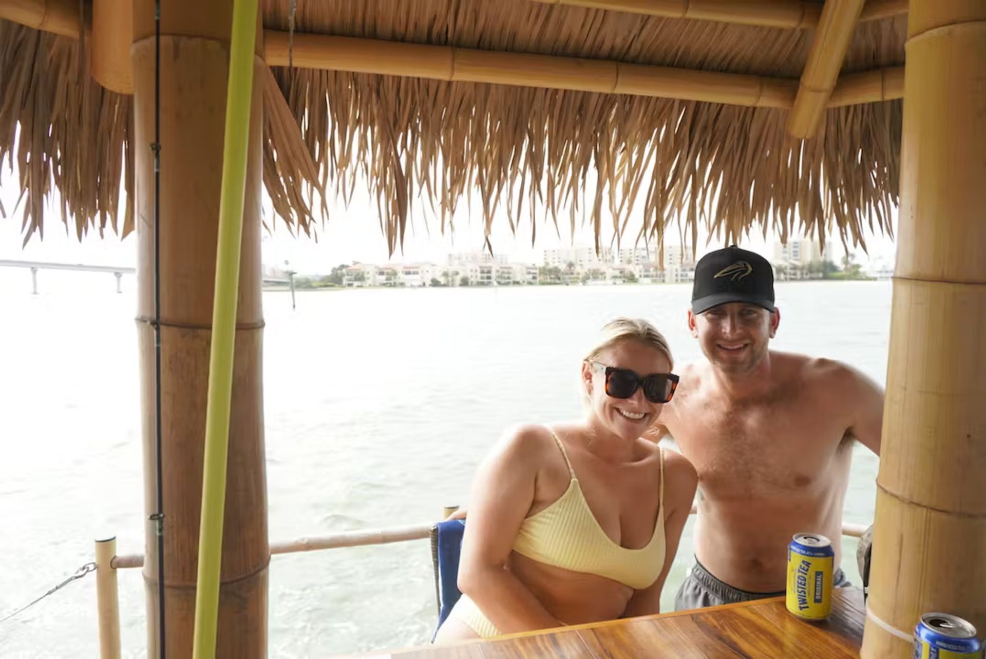 Couple in swimwear smiling under a thatched tiki hut at a waterfront bar, wooden counter with canned drinks and coastal homes and a bridge across the bay visible in the background.