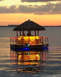 Tropical floating tiki-bar boat with a thatched roof and colorful lights reflecting on calm coastal waters at sunset.