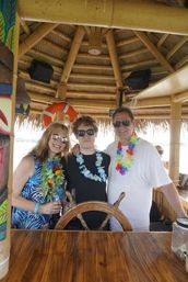 Three people in sunglasses wearing colorful Hawaiian leis pose at a wooden ship wheel inside a bamboo, thatched-roof tiki bar with a life preserver and waterfront view — tropical vacation vibe.