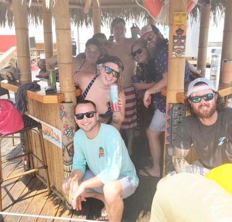 Group of friends at a beachside tiki bar on a wooden boardwalk, wearing swimsuits and sunglasses, smiling and holding colorful tumblers under a thatched roof with tropical decor.
