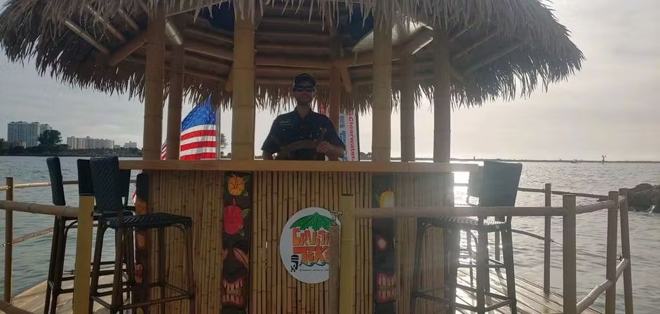 Waterfront tiki bar on a wooden pier at sunset — bamboo counter with tall stools, American flag and a silhouetted person at the helm, calm bay waters in the background.