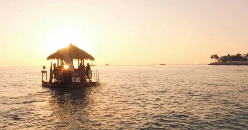 Silhouetted floating tiki raft with people enjoying a golden ocean sunset, calm water and a palm-lined coast in the distance.
