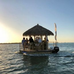 Group enjoying drinks on a tiki-style floating bar pontoon with thatched roof and outboard motor cruising coastal waters at sunset.