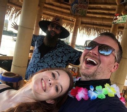 Three friends laughing in a waterfront tiki hut selfie — woman leaning in, man with sunglasses and a colorful lei, and a bearded man in a wide-brim hat waving from behind under a thatched roof with water and buildings visible beyond.