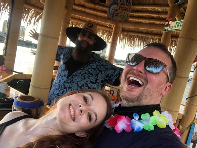 Three friends laughing in a waterfront tiki hut selfie — woman leaning in, man with sunglasses and a colorful lei, and a bearded man in a wide-brim hat waving from behind under a thatched roof with water and buildings visible beyond.