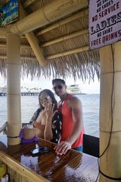 Smiling couple at an oceanfront tiki bar under a thatched roof — woman flashing a ring and holding a drink, man in a red tank top and sunglasses leaning on a shiny wooden bar, pier and palm trees over calm water in the background, vintage sign overhead.