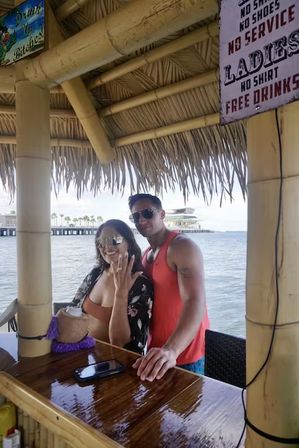 Smiling couple at an oceanfront tiki bar under a thatched roof — woman flashing a ring and holding a drink, man in a red tank top and sunglasses leaning on a shiny wooden bar, pier and palm trees over calm water in the background, vintage sign overhead.