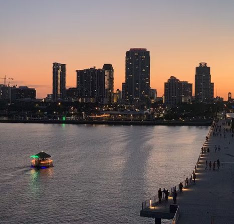 Silhouetted city skyline at sunset over a calm waterfront, small illuminated boat on the water and a busy pier promenade with people walking.