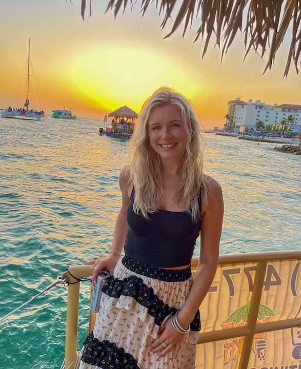 Smiling woman on a seaside railing at a tropical oceanfront sunset, turquoise water with boats and a thatched tiki bar, beachfront resort in the background